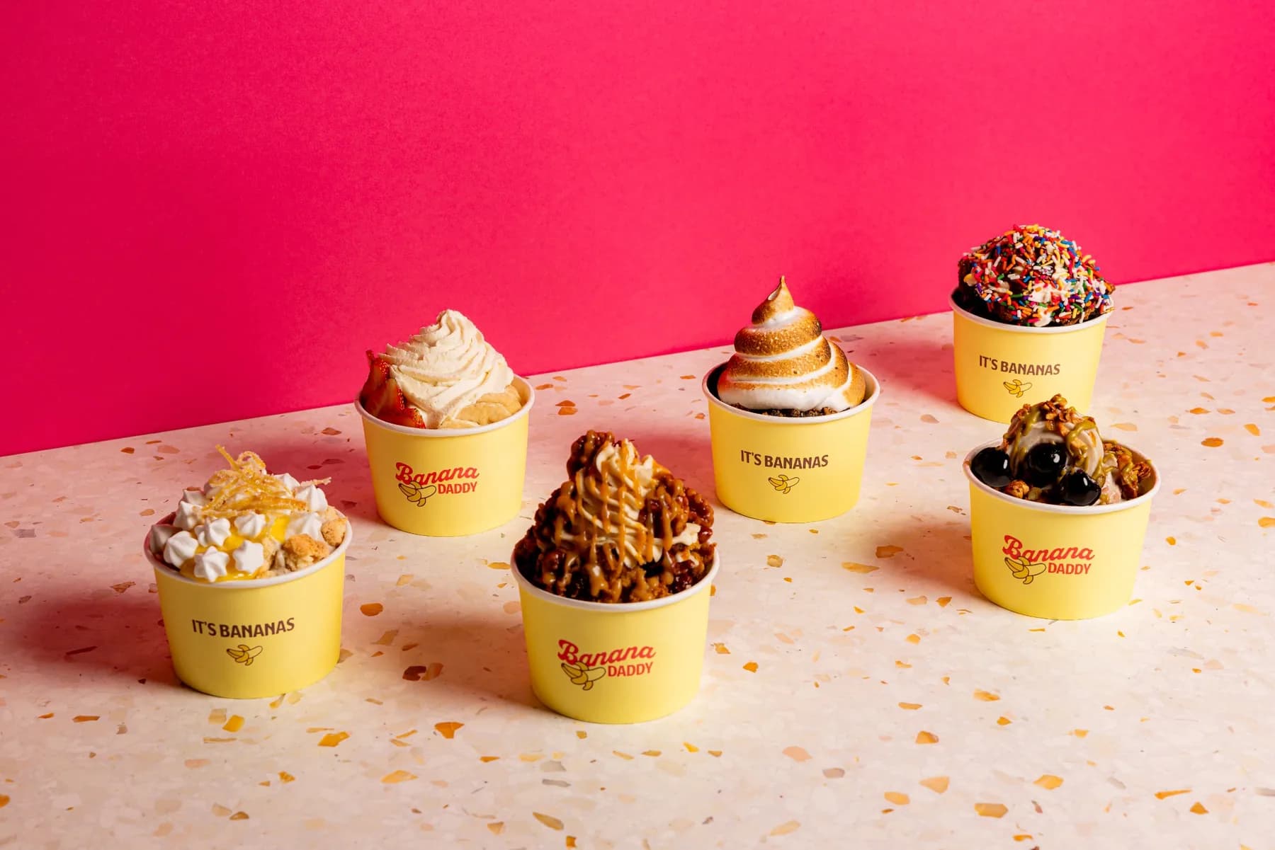 Six soft serve sundaes in yellow Banana Daddy cups on a terrazzo counter.